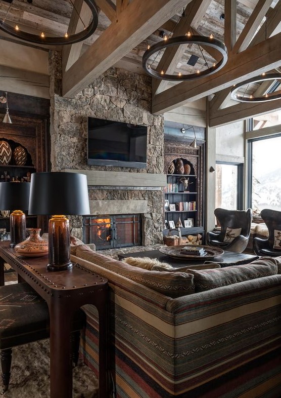 a stylish chalet living room clad with wood, with a stone fireplace, boho and modern furniture and round chandeliers