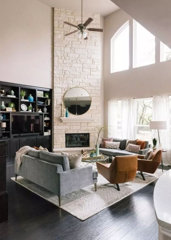 a stylish modern living room with a white faux stone corner fireplace with a small mantel, a round mirror and double-height windows