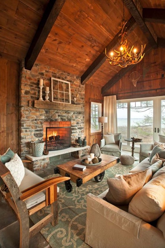 a welcoming chalet living room with a wooden ceiling and walls, a stone clad fireplace, modern and rustic furniture and an antler chandelier