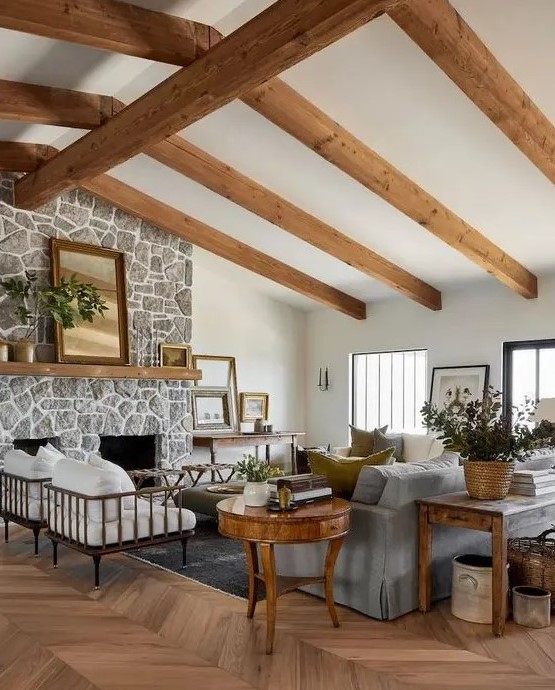 a welcoming modern farmhouse living room with wooden beams, a fireplace clad with stone, a grey sofa and white chairs, stained and refined furniture