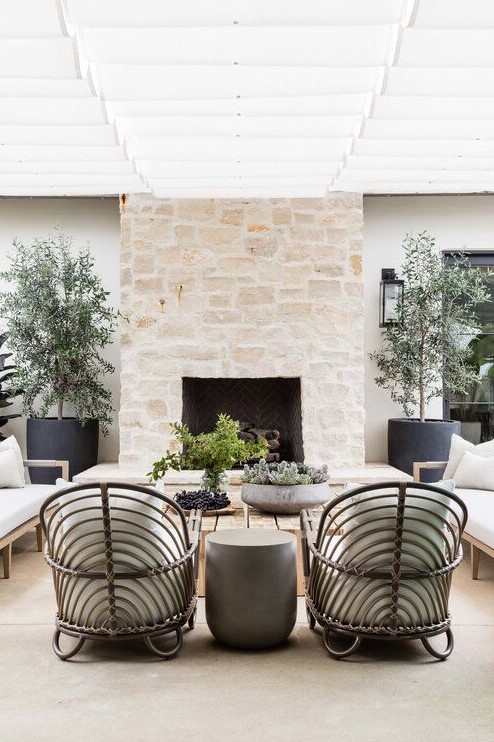 an inviting modern terrace with a large stone clad fireplace, rattan and wood furniture, potted greenery and trees is chic