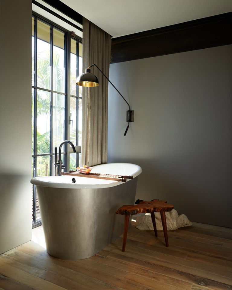 a cool oval free standing tub is perfect for any interesting bathroom design The bathroom is clean and features a full height window with a curtain, a silver oval tub and a catchy wall lamp