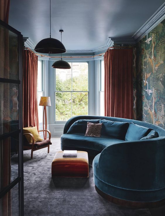 a moody and bold living room with a curved blue sofa, orange textiles and moody bird print wallpaper on one wall