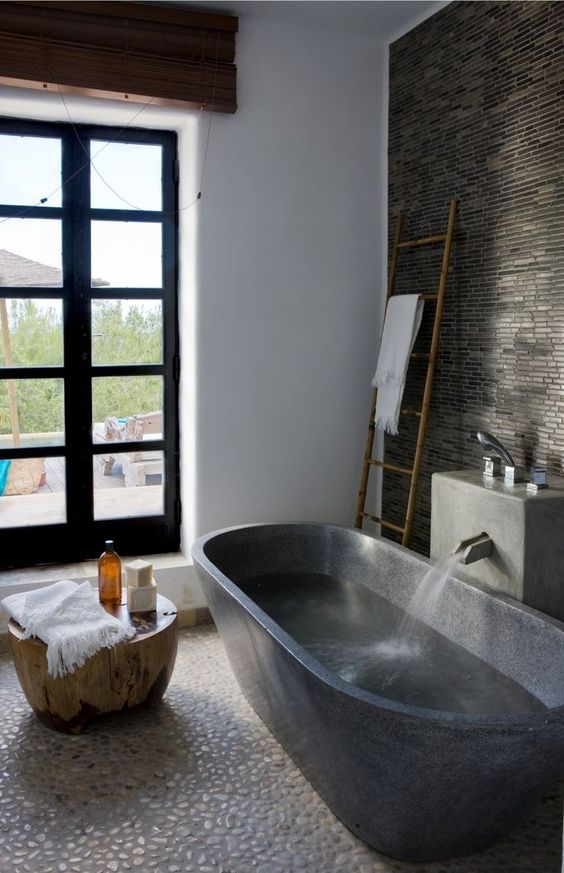 a moody bathroom with a white pebble tile, dark stone on the wall and a dark stone bathtub plus a wooden side table