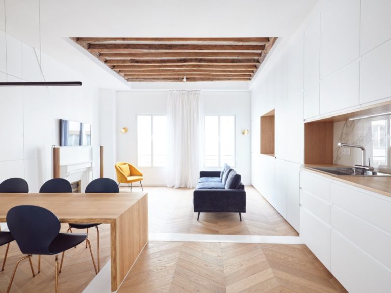 The living room is done with a faux fireplace, a blue sofa, a yellow chair and airy and light curtains, wooden beams on the ceiling add coziness