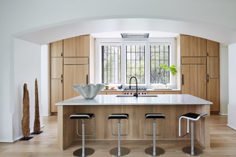 The kitchen is done with light colored wooden cabinets and a kitchen island plus a large window instead of a backsplash The kitchen is done with light-colored wooden cabinets and a kitchen island plus a large window instead of a backsplash