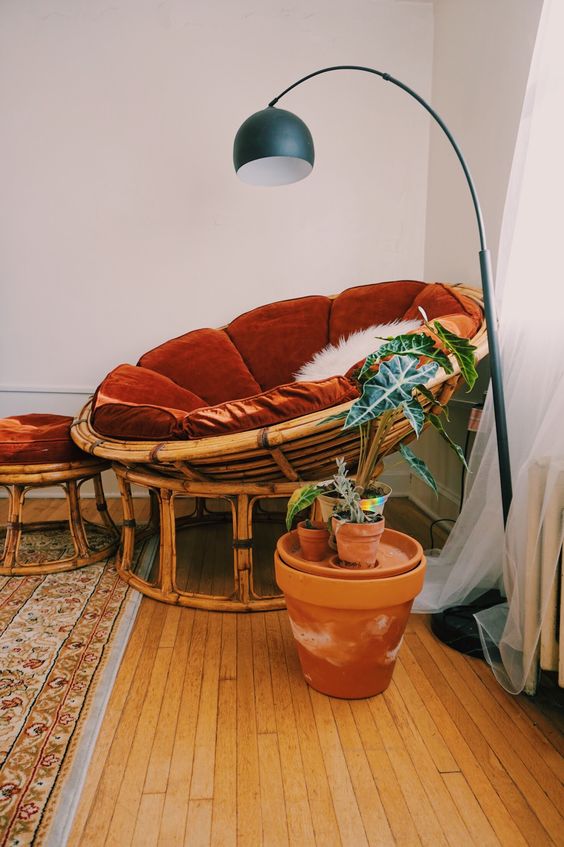 a rattan papasan chair with a rust velvet futon and a matching footrest plus some potted plants