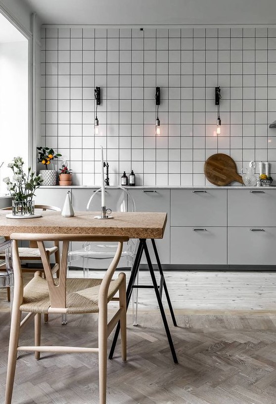 a Nordic kitchen with grey cabinets, a white tile backsplash, sconces that hold bulbs, a table with a cork countertop and wooden chairs