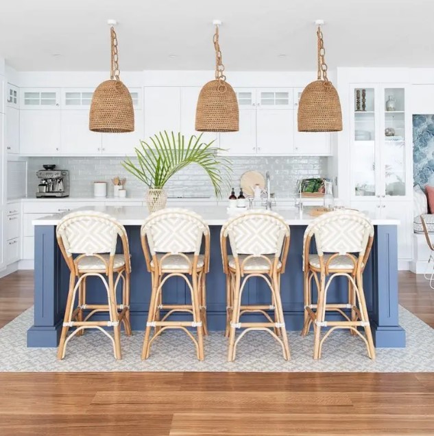 a beach kitchen with white cabinetry, a navy kitchen island, aqua green tiles on the backsplash rattan chairs, woven lamps and greenery