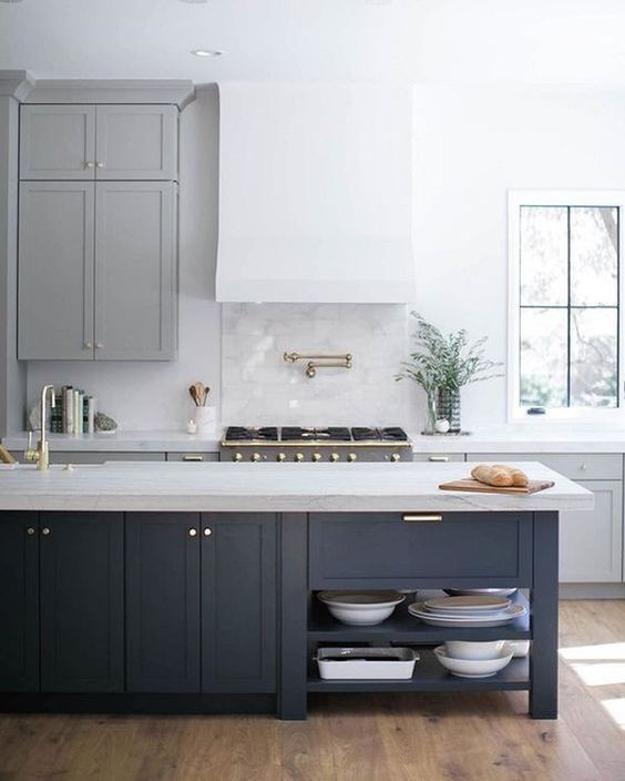 a beautiful and airy contemporary kitchen with grey cabinets, a white hood, a navy kitchen island with a stone countertop and gold touches