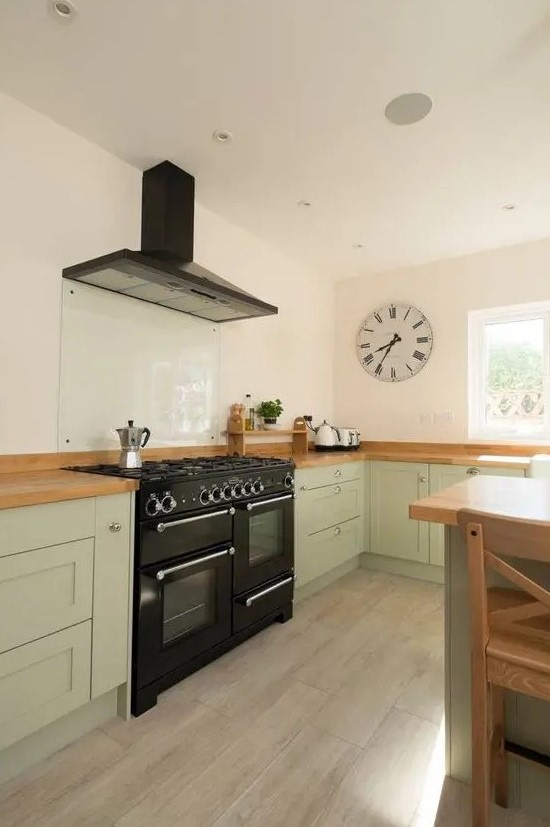 a beautiful sage green kitchen with butcherblock countertops, a black cooker and a hood, a glass backsplash is chic