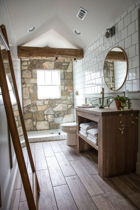 a chalet bathroom with a stone wall, a wooden floor and a wooden vanity, a couple of mirrors and a wooden beam