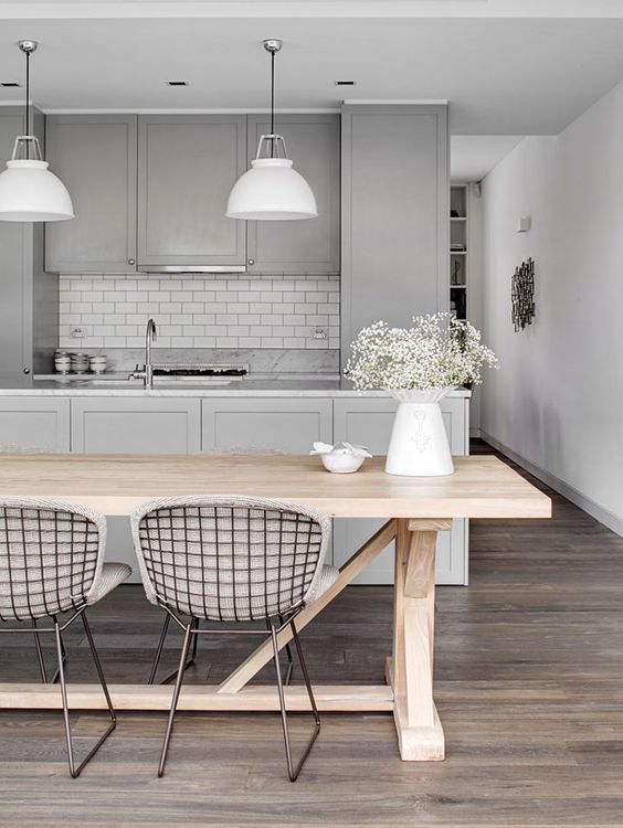 a chic kitchen with grey shaker cabinets, a white subway tile backsplash, white pendant lamps, a stained table and metal wire chairs