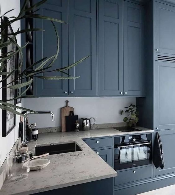 a chic kitchen with navy cabinets, neutral stone countertops, stainless steel fixtures and greenery