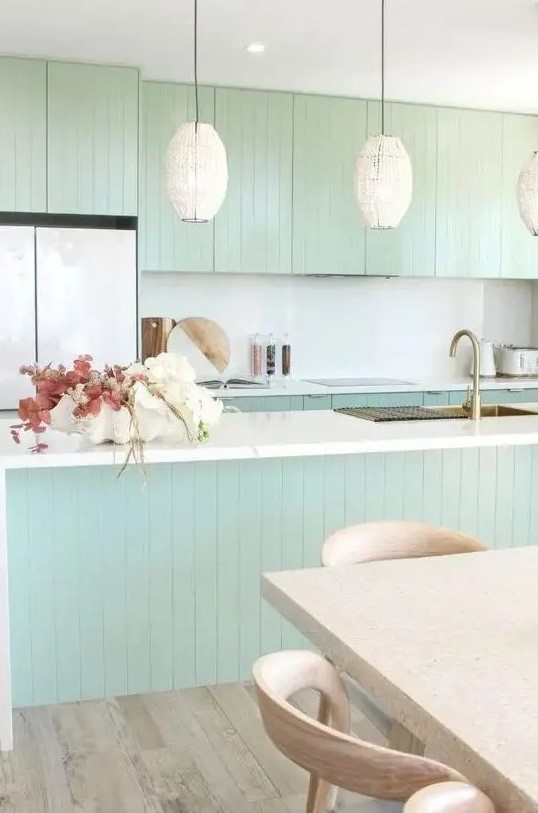 a chic mint green slatted kitchen with a white backsplash and countertops, cool pendant lamps, a terrazzo table and stained chairs