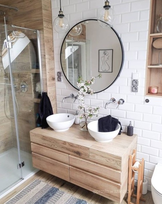 a contemporary bahtroom clad with white subway tiles, wood look tiles in the shower, a wooden vanity, round sinks and a round mirror