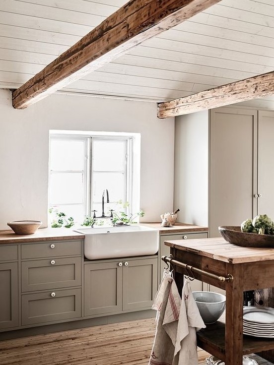 a cozy farmhouse neutral kitchen with grey cbainets, wooden countertops and beams, a wooden cart kitchen island