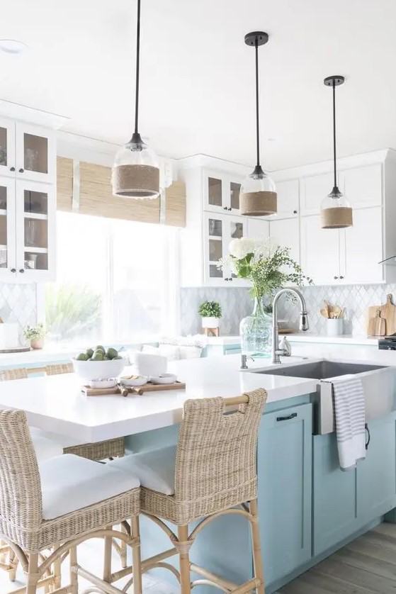a gorgeous beach kitchen with white shaker cabinets, a geo tile backsplash, woven blinds, a light blue kitchen island and woven stools plus glass lamps with burlap touches