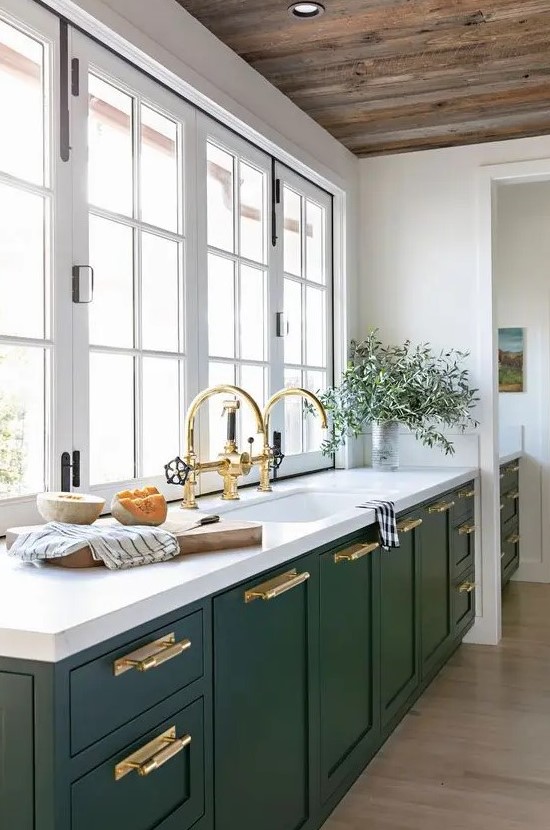 a hunter green modern kitchen with white stone countertops, gold handles and fixtures and a window instead of a backsplash