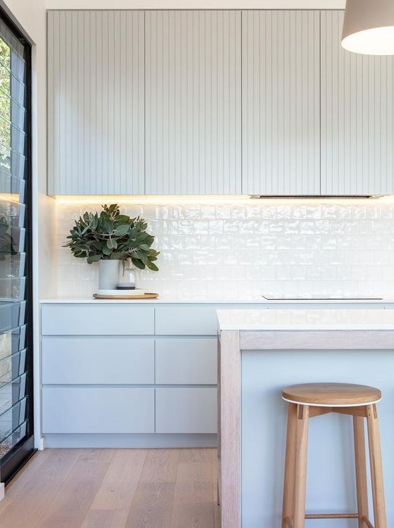 a minimalist coastal kitchen with very light blue and off-white cabinets, a white tile backsplash, a kitchen island and wooden stools