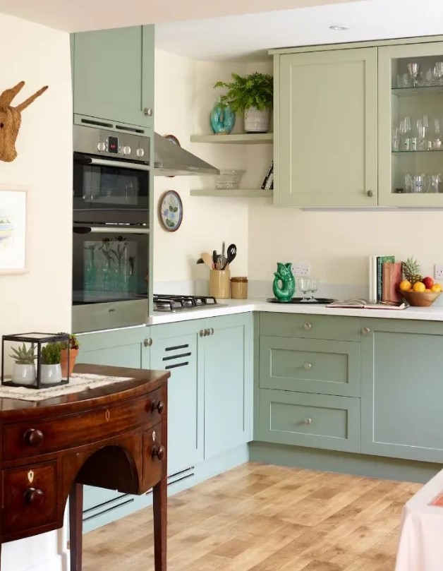 a modern sage green kitchen with shaker cabinets and glass front ones, a white backsplash and countertops plus a vintage sideboard