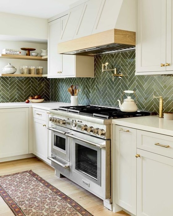 a refined kitchen with creamy cabinets, a green herringbone tile backsplash, a white hood and gold sconces