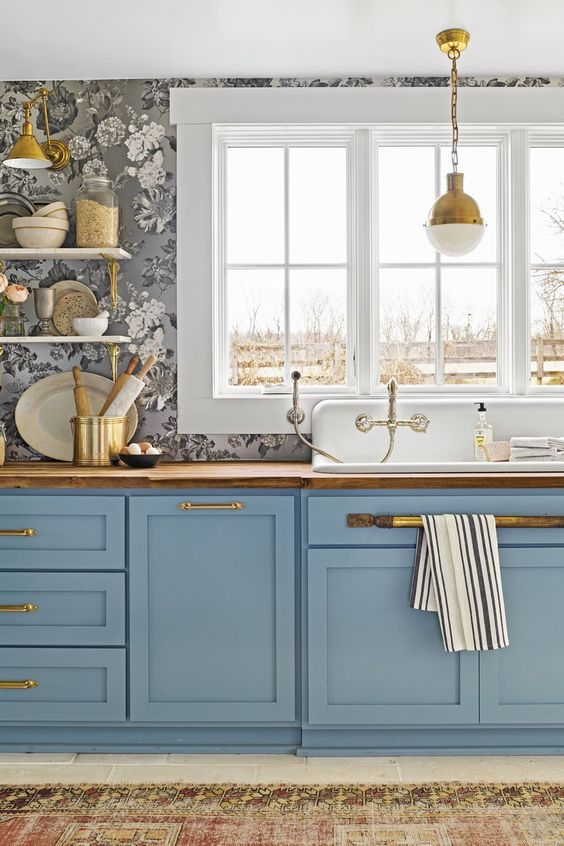 a refined kitchen with grey floral print wallpaper, blue shaker cabinets, butcherblock countertops, gold handles and a gold pendant lamp