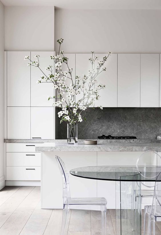 a refined minimalist kitchen done in white, with a grey stone backsplash and countertops, a glass table and acrylic chairs