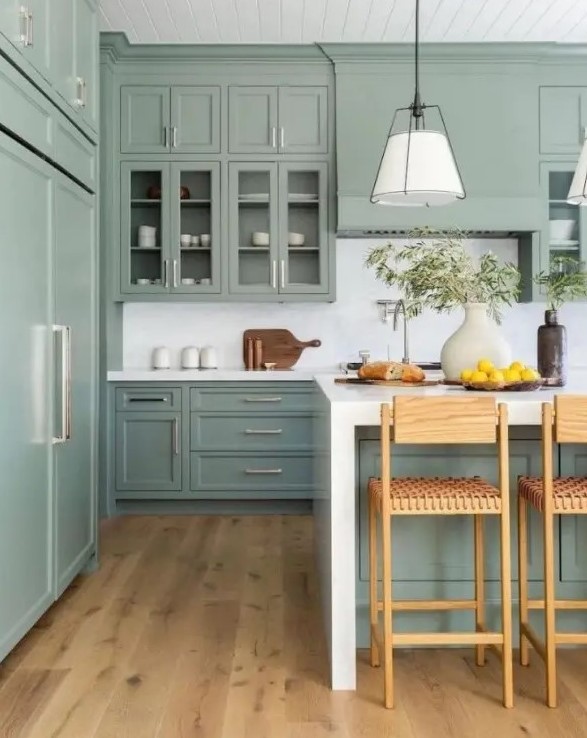 a sage green kitchen with shaker and glass front cabinets, white stone countertops and a backsplash, woven stools