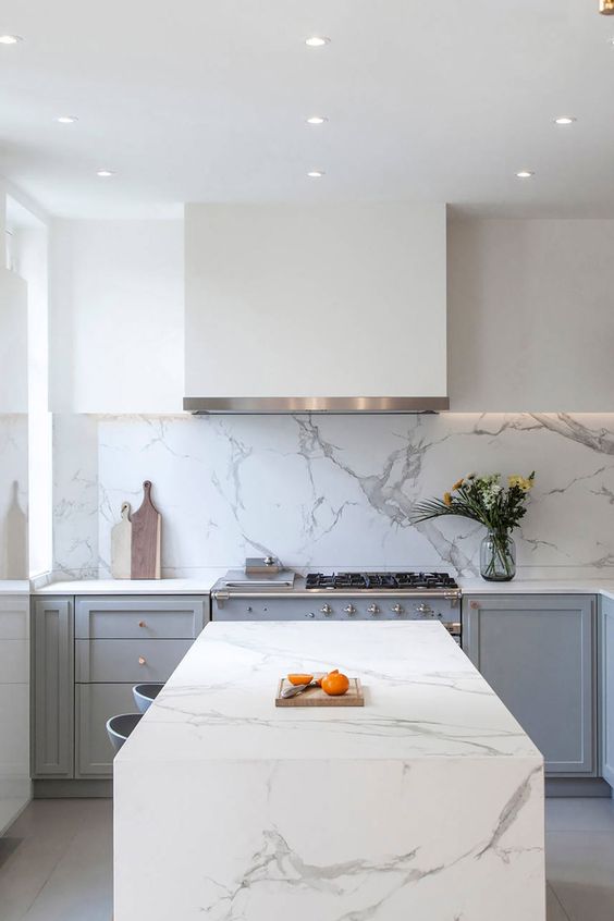 a serene grey and white kitchen with only a lower row of cabinets in grey, a white hood, a white marble backsplash and a matching kitchen island