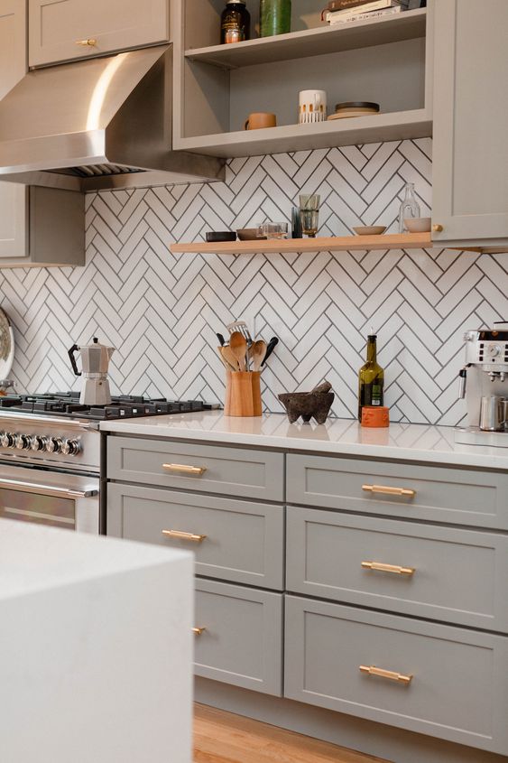 a sophisticated light grey kitchen with shaker cabinets, a white chevron tile backsplash, white countertops and a shiny metal hood