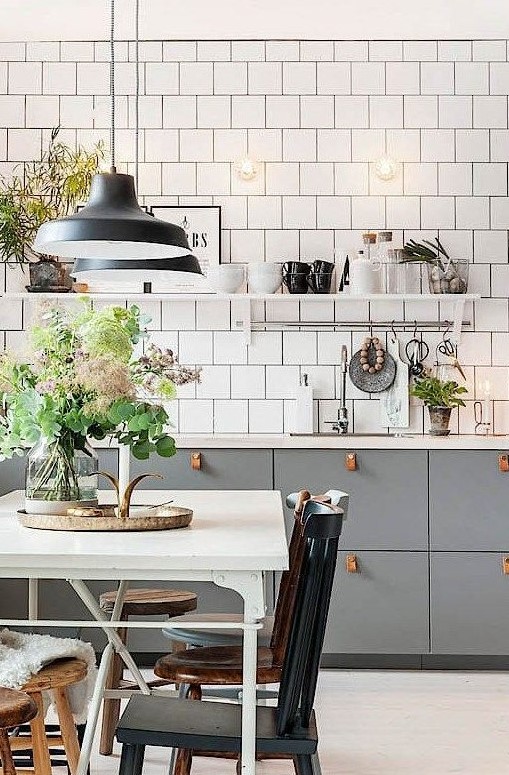 a stylish Scandinavian kitchen with grey cabinets and leather pulls, white square tiles, vintage chairs and pendant lamps