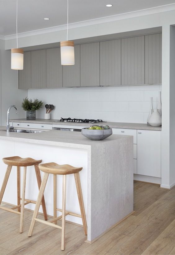 a stylish minimalist kitchen with white cabinets and grey upper ones, a white tile backsplash, a kitchen island with a waterfall countertop and pendant lamps