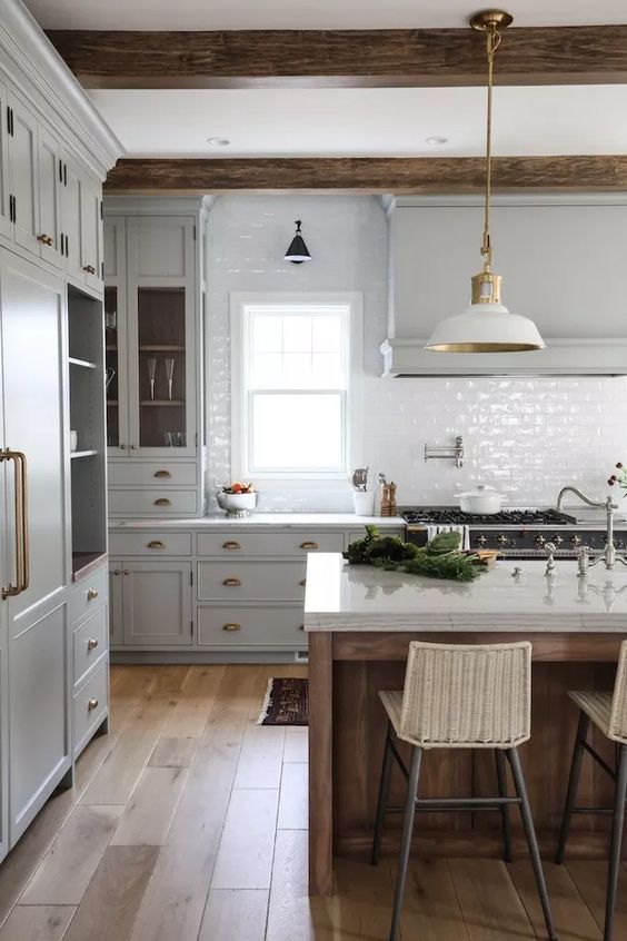 a vintage kitchen with dove grey cabinets, white tile backsplash, white lamps and wooden beams plus a kitchen island