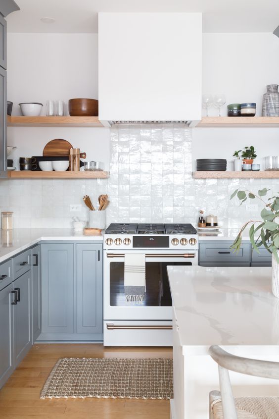 an airy kitchen with light blue cabinets, a white Zellige tile backsplsh, open shelves and white countertops