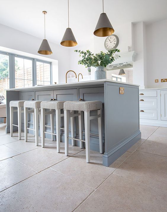 an airy kitchen with pale blue cabinets, a blue kitchen island, grey pendant lamps and creamy stools