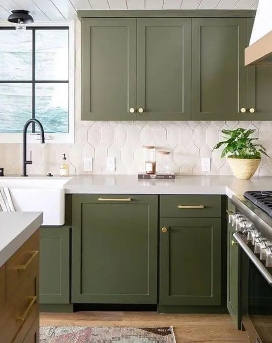 an olive green farmhouse kitchen with shaker cabinets, a catchy white tile backsplash, black fixtures and gold handles and knobs