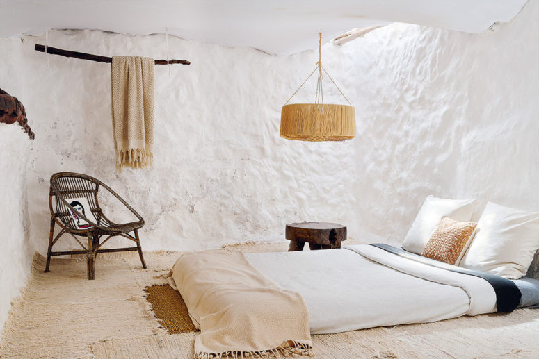 The master bedroom shows off layered rugs, a mattress, a wicker chair, a wooden stool, a wicker lamp and a lanket hanging on a branch