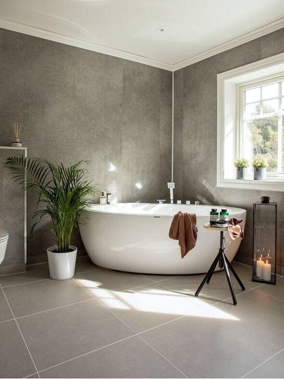 a beautiful grey bathroom with two kinds of tiles, potted plants, a free-standing tub, a candle lantern and a table