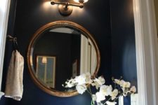 a chic powder room with navy walls, a stenciled gold and white ceiling, a gold frame mirror, a sconce and a stone sink