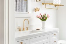 a chic white bathroom with gold fixtures, knobs, a shelf and sconces is a pretty space