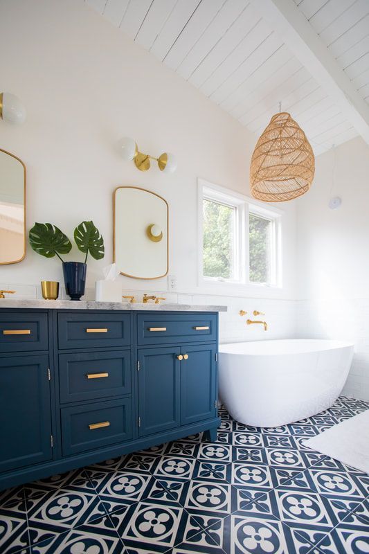a coastal bathroom with blue and white printed tiles, a blue and gold vanity, gold frame mirrors, a woven lamp and a free-standing tub