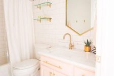 a contemporary bathroom clad with white tiles, with a light pink vanity, gold fixtures, knobs and a geometric mirror