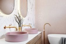 a contemporary bathroom with blush hex tiles on the wall, pink sinks, glam fixtures and lit up gold frame mirrors