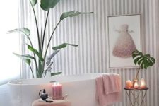 a contemporary glam bathroom with a striped wallpaper wall, a pink dress artwork, pink towels and an ottoman, a pink rug and candles plus touches of gold