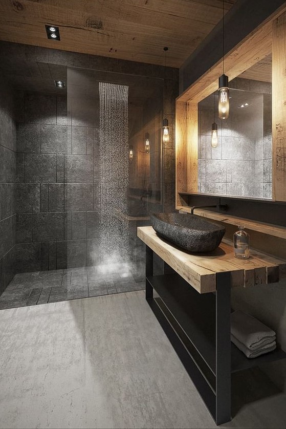 a creative grey bathroom done with grey tiles, a wood and metal vanity, a stone sink and a large lit up mirror
