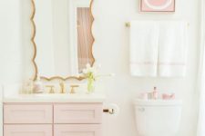 a glam contemporary bathroom with a light pink vanity, artwork and accessories, gold fixtures and a a catchy mirror in a gold frame
