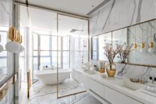 a glam white bathroom all done with marble, with pendant gold lamps, gold frames and a mirror in a gold frame