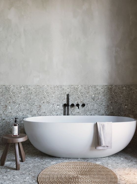 a grey bathroom done with stone and plaster, with a free-standing tub, a wooden stool and a jute rug