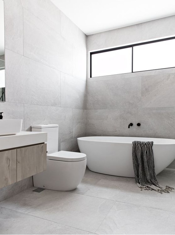 a grey minimalist bathroom clad with large scale tiles, an oval tub, a long and narrow window and a floating vanity
